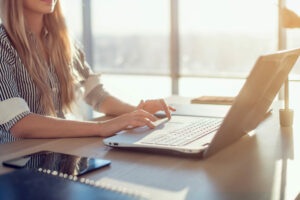 A blond woman sitting at a computer desk typing on a laptop.