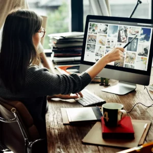 A woman sitting at a desk, facing a computer monitor. She is pointing at something on the monitor with a pencil.