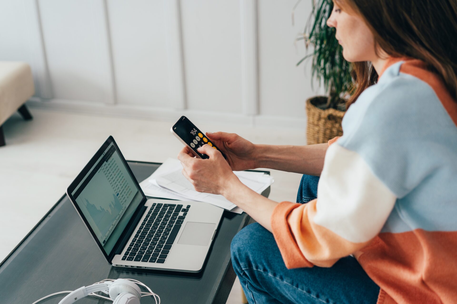 A woman sitting on a couch, holding a calculator. On a low table in front of her is an open laptop.