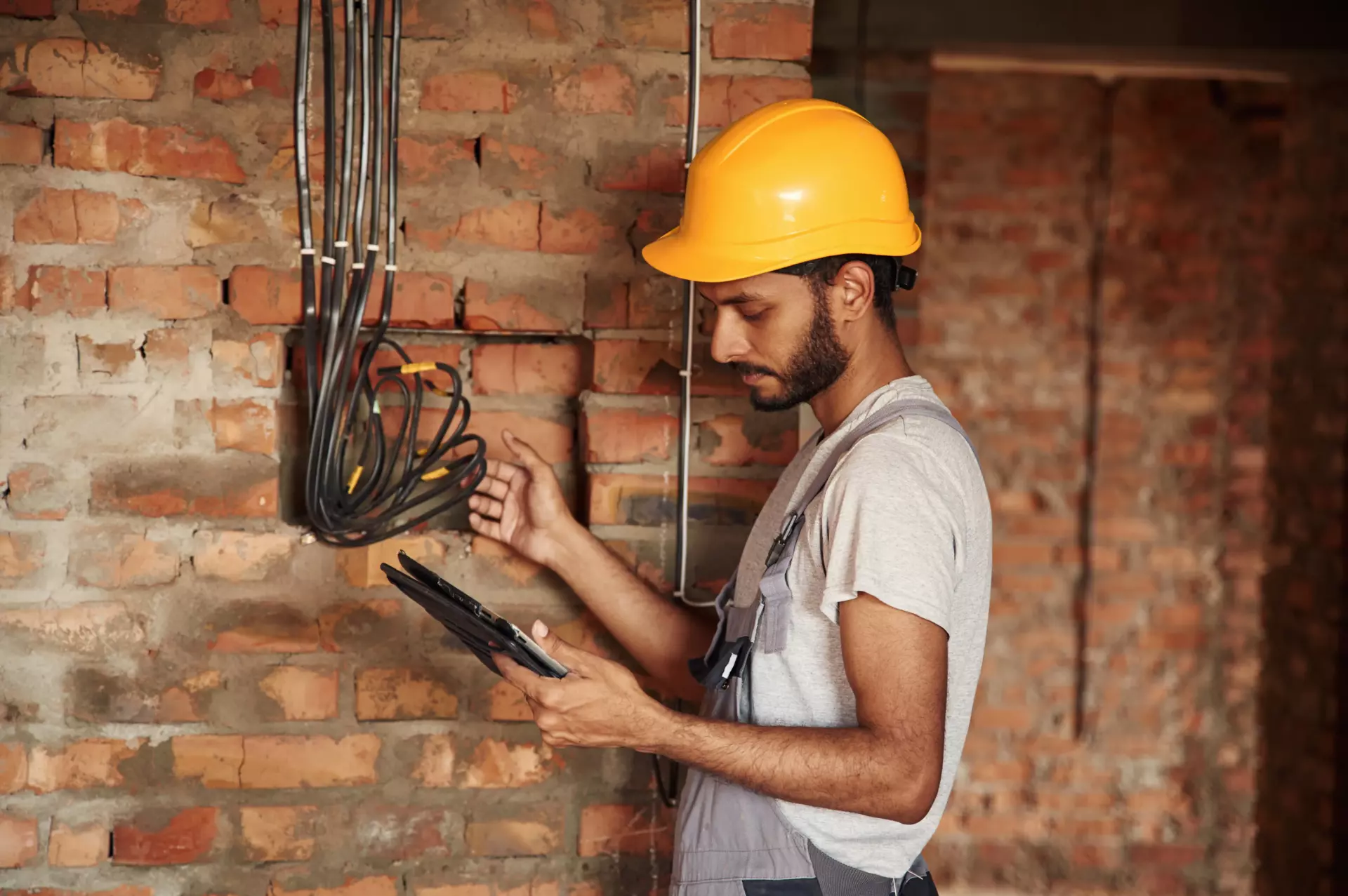 A man wearing a hard hat holds a tablet in one hand and points to some wires in a brick wall with the other.