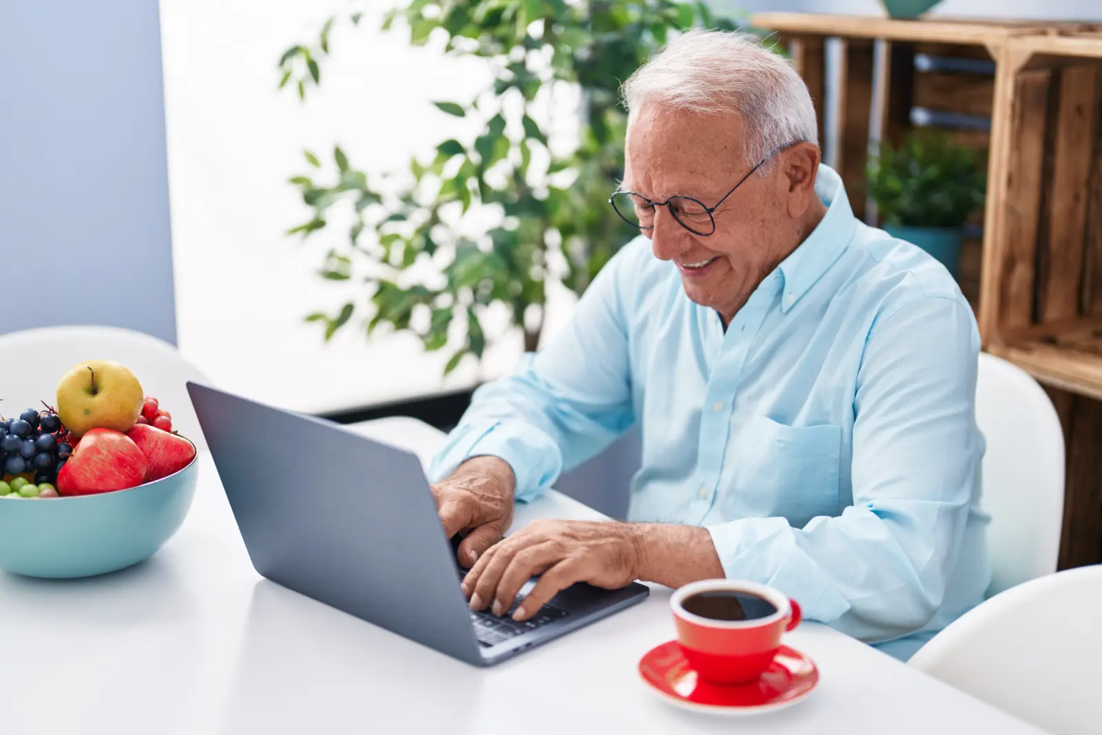 An elderly man wearing glasses is sitting at a table, looking at an open laptop.