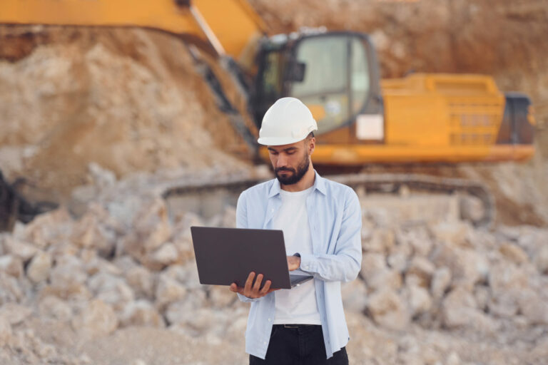 A man wearing a hard hat is holding an open laptop. Behind him is an excavator.