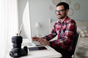 A man in a red flannel shirt sits at a computer with his hands on the keyboard.